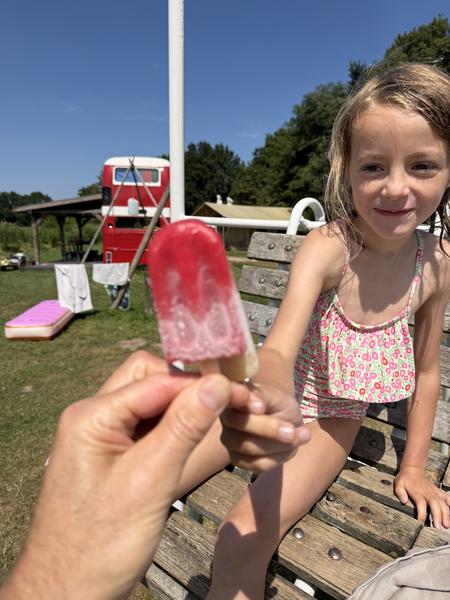A young child with a bright smile is about to receive a red popsicle from an unseen person, sitting on a wooden bench under a clear blue sky